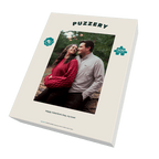A boxed jigsaw puzzle package shown at an angle on a black background. The front of the box is cream-colored with the brand name 'PUZZERY' at the top, a photo of a couple in the center, a teal puzzle-piece icon with '500' on the right, and the phrase 'Happy Valentine's Day, my love!' near the bottom.