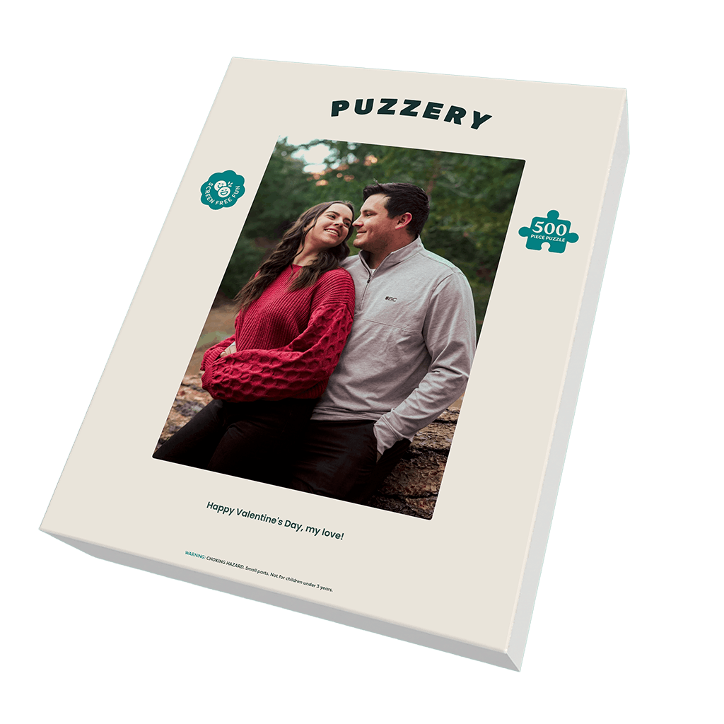 A boxed jigsaw puzzle package shown at an angle on a black background. The front of the box is cream-colored with the brand name 'PUZZERY' at the top, a photo of a couple in the center, a teal puzzle-piece icon with '500' on the right, and the phrase 'Happy Valentine's Day, my love!' near the bottom.